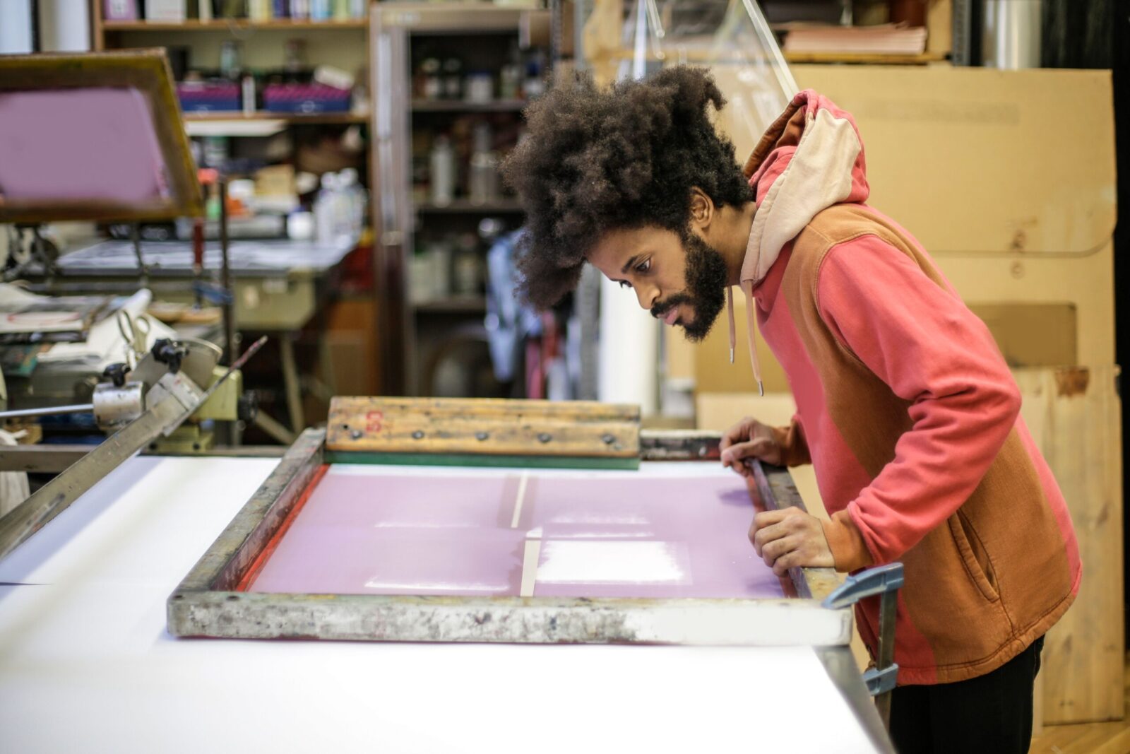 Side view of focused young bearded ethnic craftsman in casual hoodie standing near workbench with wooden frame and preparing colorful material in modern workshop